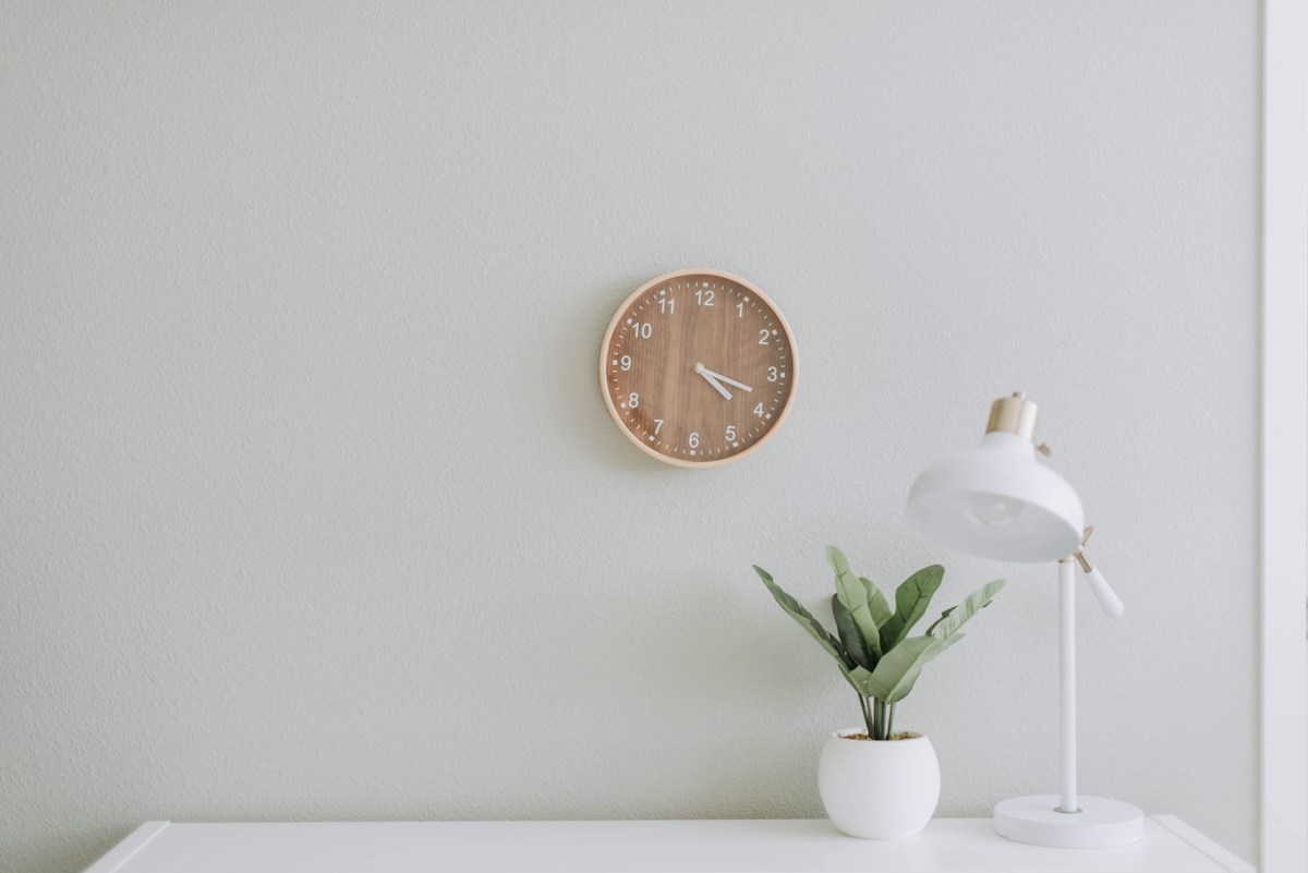 A white desk with a reading lamp, small plant and wooden wall clock